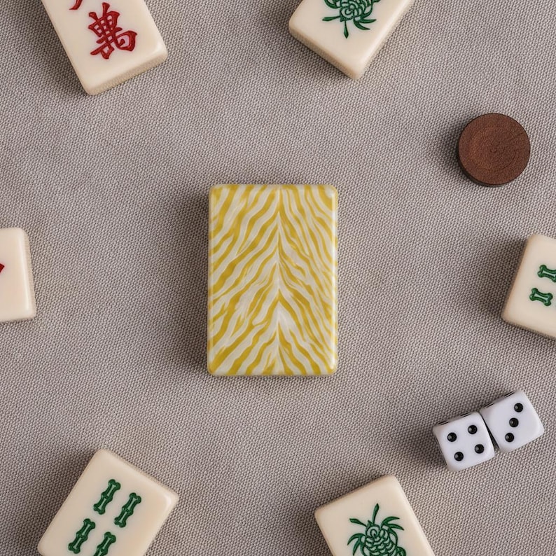 May include: A rectangular mahjong tile featuring a yellow and white tiger stripe pattern on its back, surrounded by several standard off-white mahjong tiles with green and red markings, a small brown circular token, and two white six-sided dice.
