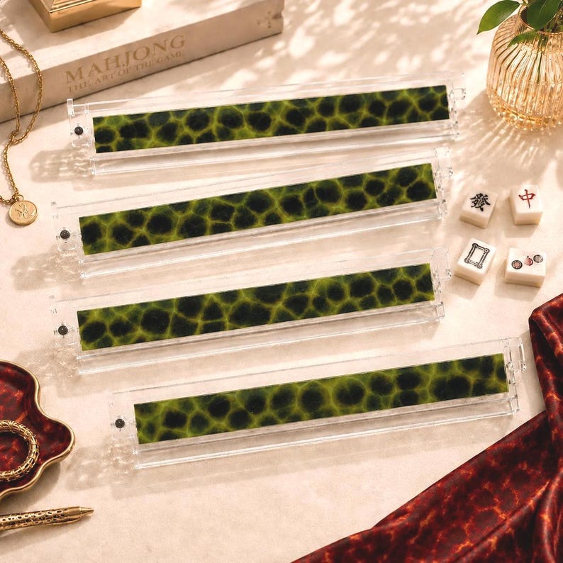 May include: Four clear acrylic mahjong tile racks, each featuring a long, horizontal insert with a dark green and black mottled pattern. The racks are arranged in a parallel stack, showcasing their transparent construction and textured green inlay.