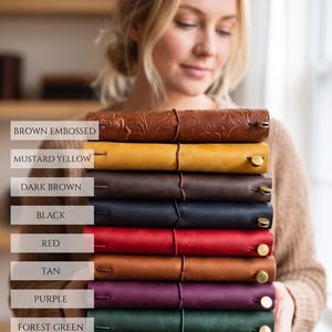May include: A stack of leather journals in various colors, including brown embossed, mustard yellow, dark brown, black, red, tan, purple, forest green, and teal blue. Each journal is secured with a leather strap and a brass button closure.