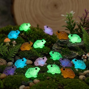 May include: A collection of glowing miniature frog figurines in various colors, including green, blue, orange, and purple. The frogs are arranged on a bed of green moss, creating a whimsical fairy garden scene. Some frogs have a dotted pattern.