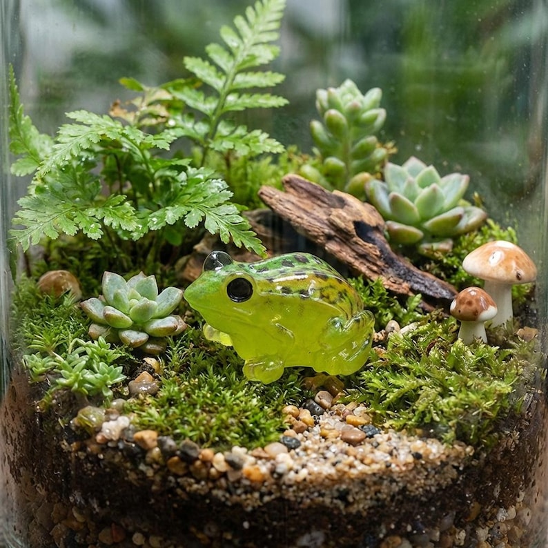 May include: A miniature terrarium in a glass jar, featuring a vibrant green frog figurine, small mushrooms, succulents, and lush green moss. The scene includes small pebbles and a piece of driftwood, creating a miniature garden scene.