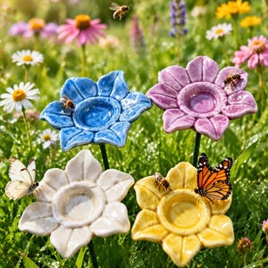 Puede incluir: Cuatro estacas de jardín de cerámica con forma de flor en azul, morado, blanco y amarillo. Cada pieza presenta un cuenco central rodeado por seis pétalos, montado sobre un tallo verde fino, exhibido entre flores silvestres e insectos.