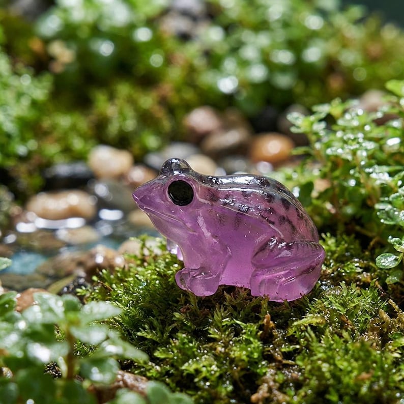 May include: A small, translucent purple frog figurine with black eyes sits on a bed of green moss. The frog's body has a slightly iridescent quality, and the background features small stones and more greenery.