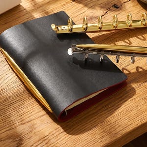 May include: A black leather-bound notebook with gold-colored accents and a gold pen. The notebook is open, revealing red-edged pages. A gold-colored binder mechanism is also visible. The items are on a wooden surface.