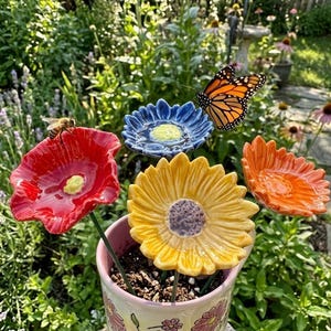 Puede incluir: Un conjunto de cuatro estacas de jardín de cerámica con forma de flor en rojo, azul, amarillo y naranja, cada una con un centro texturizado, dispuestas en una maceta de cerámica clara, con una mariposa monarca en la flor azul y una abeja en la roja.