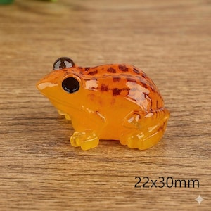 May include: A small, orange frog figurine with dark brown spots and black eyes. The frog is made of a translucent material and measures 22mm x 30mm. The figurine is sitting on a wooden surface.
