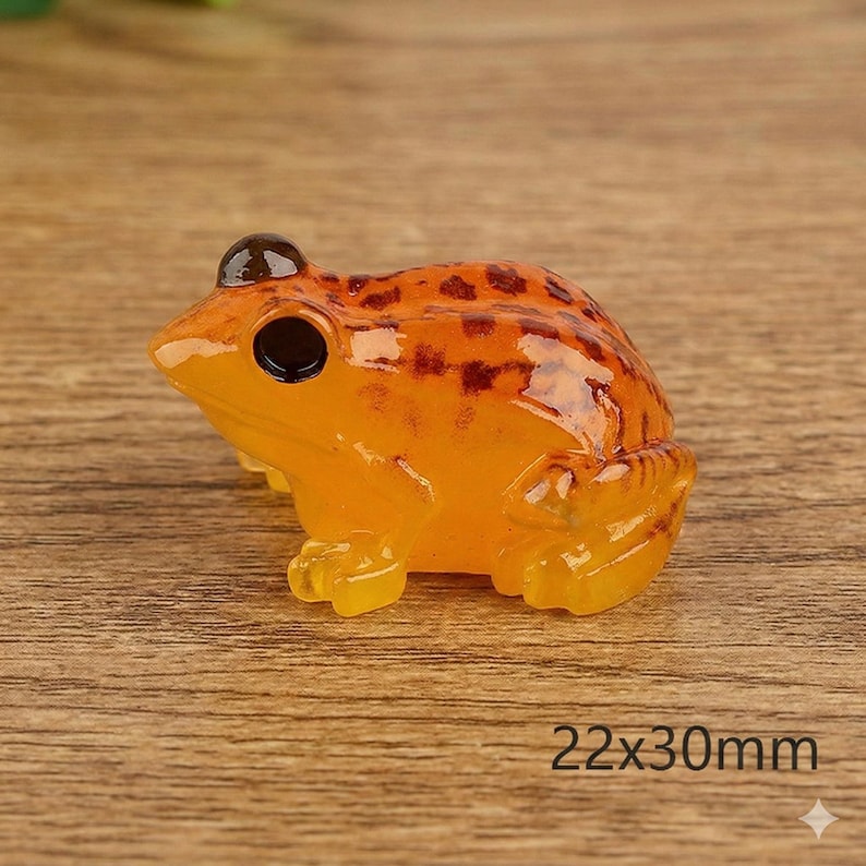 May include: A small, orange frog figurine with dark brown spots and black eyes. The frog is made of a translucent material and measures 22mm x 30mm. The figurine is sitting on a wooden surface.