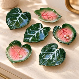 May include: Six ceramic trinket dishes shaped like leaves. Three dishes feature deep green surfaces with white vein details, while the other three display a gradient of green edges transitioning to pink centres with prominent red vein markings.