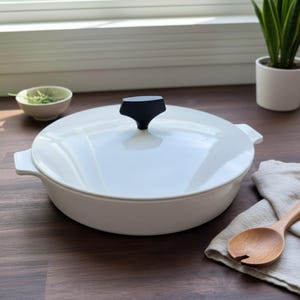 May include: A white ceramic casserole dish with a black handle on the lid. The dish is on a wooden surface, with a small bowl of herbs and a potted plant in the background. A wooden spoon rests on a linen cloth.