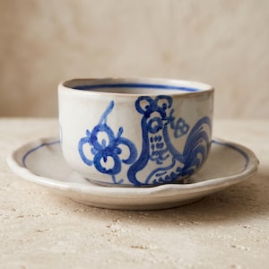 May include: A ceramic tea cup and matching saucer featuring a hand-painted blue rooster and floral motif on an off-white glazed surface. The cup has a straight rim and the saucer has a slightly scalloped edge with a thin blue painted line.