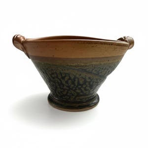 May include: A handcrafted ceramic bowl with a wide, flared shape. The bowl features a textured, dark green and brown exterior with two small handles. The rim is a lighter shade of brown. The bowl sits on a small, round base.