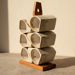 May include: A set of six off-white ceramic mugs with a unique irregular shape and a drip glaze finish, stacked in two columns on a wooden stand. The stand features a central vertical handle with a circular cutout at the top for easy carrying.