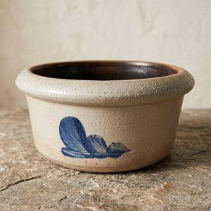 May include: A round stoneware bowl with a textured beige exterior and a dark brown interior. The bowl features a thick, rounded rim and a hand-painted blue floral motif on the side, showcasing a rustic, speckled finish throughout the clay surface.