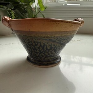 May include: A handcrafted ceramic bowl with a wide, flared shape. The bowl features a textured, dark blue lower section, transitioning to a brown and tan upper rim. Two decorative handles are on either side. The bowl is approximately 10 inches in diameter.