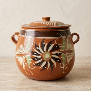 May include: A round terracotta pot with a matching lid and two side handles. The exterior features a dark brown band near the top and a hand-painted floral design in black, white, and green, with swirling white lines and fern-like leaf patterns.
