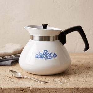 May include: A white teapot with a blue floral pattern on the front, featuring a black handle, a black lid knob, and a silver-toned metal band around the neck. The teapot has a rounded body and a small spout, accompanied by a silver-toned spoon.
