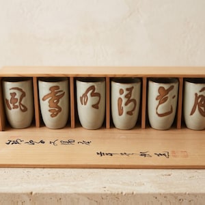 May include: A set of six light gray ceramic cups with brown calligraphic characters, arranged in a light-colored wooden storage box. The front of the box features black ink calligraphy and a small red seal stamp on the bottom right corner.