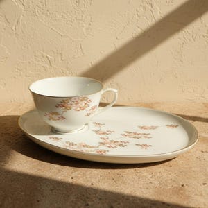 May include: A white porcelain teacup and matching oval saucer set featuring a delicate floral pattern in shades of brown, gold, and green. Both pieces are finished with a thin gold rim, showcasing a refined design with a curved handle.