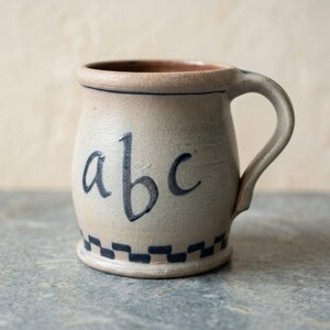May include: A beige ceramic mug with a rounded body and a single handle. The front features the lowercase letters abc painted in dark blue. A dark blue checkered pattern encircles the base of the mug, and a thin blue line circles the upper rim.