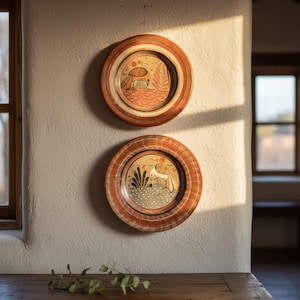 May include: Two decorative plates with a rustic design. The plates feature a reddish-brown rim with a patterned border. The center of each plate displays a hand-painted scene with a bird and foliage in shades of orange, beige, and brown. They are mounted on a white wall.