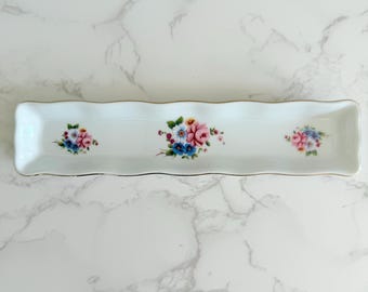 May include: A rectangular white ceramic serving dish with a scalloped edge and gold trim. The dish is decorated with three floral bouquets in shades of pink, blue, and yellow. A decorative piece for serving appetizers or desserts.