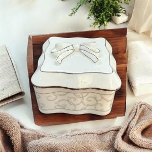 May include: A white ceramic jewellery box with a bow-shaped lid, accented with silver trim. The box sits on a wooden tray and features a decorative scroll pattern on the lower section. The design is elegant and suitable for storing small items.