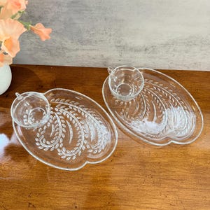 May include: Two clear glass teacup and saucer sets. The oval saucers feature a leaf design and a circular indentation for the teacup. The teacups have a handle and are also clear glass. The sets are on a wooden surface.