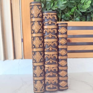 Könnte beinhalten: Drei dekorative Buchstützen mit dunkelbraunem Grund und goldfarbenen, verzierten Designs. Die Buchstützen weisen verschiedene Muster auf, darunter Kreuze, Rankenwerk und Rauten. Sie sind vertikal angeordnet und suggerieren eine klassische oder Vintage-Ästhetik.