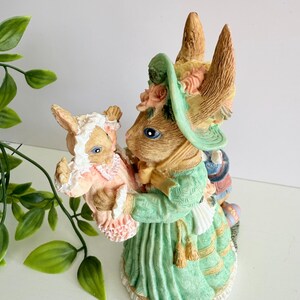 Puede incluir: Figura decorativa de Pascua que representa una coneja madre sosteniendo un conejito bebé. La madre lleva un vestido verde y un sombrero adornado con flores. El conejito bebé viste un traje rosa. La figura está pintada a mano con detalles.