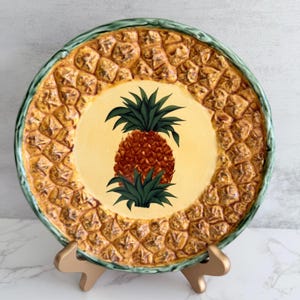 May include: A decorative ceramic plate with a pineapple design. The plate has a textured, yellow and brown exterior and a green rim. The centre of the plate has a yellow background with a detailed pineapple illustration.
