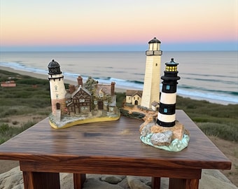 May include: Three decorative lighthouse figurines on a wooden table. One is white and black striped, another is white, and the third is a cottage-style lighthouse. The background shows a beach and ocean under a colorful sky.