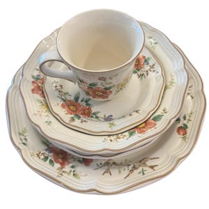 May include: A cream-colored, floral-patterned dish set featuring a teacup, saucer, and two plates. The set has a scalloped edge and is decorated with red, yellow, and green flowers, and small birds. The set is designed for serving tea or a meal.