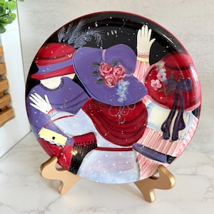 May include: A decorative plate with a painted design of women in hats. The plate has a black background with red and white accents. The women's hats are purple and red, with floral and ribbon details. The plate is displayed on a gold-coloured stand.