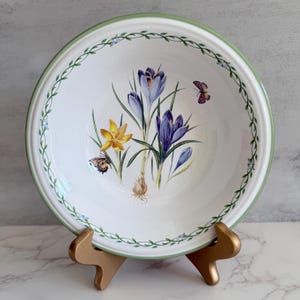 May include: White ceramic bowl with a green rim and a floral design featuring purple and yellow crocuses and butterflies. The bowl is supported by a gold-colored stand. A green vine pattern decorates the rim.
