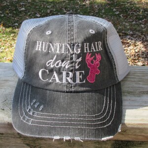 May include: A grey and white trucker cap featuring the embroidered text "HUNTING HAIR don't CARE" in white. A pink glitter deer head graphic is on the right. The cap has a distressed brim.
