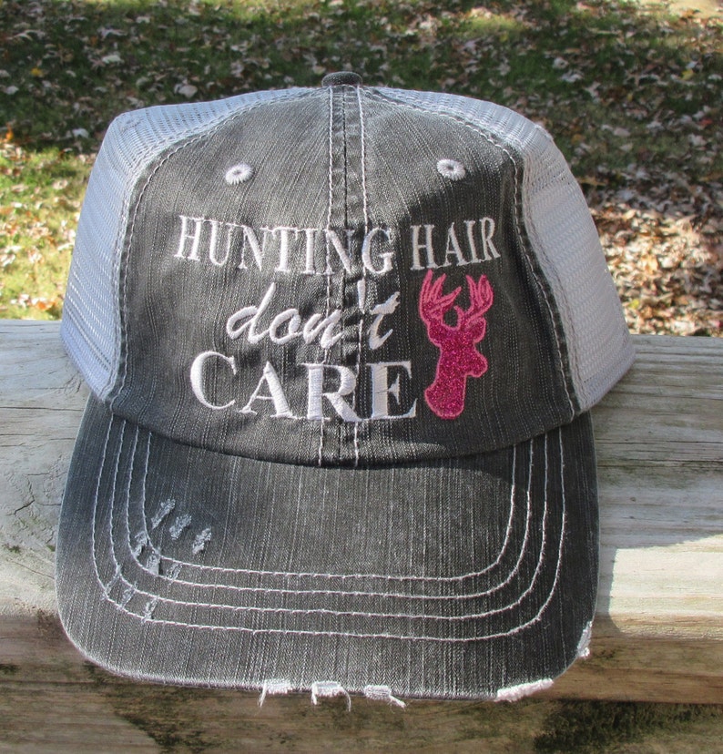 May include: A grey and white trucker cap featuring the embroidered text "HUNTING HAIR don't CARE" in white. A pink glitter deer head graphic is on the right. The cap has a distressed brim.