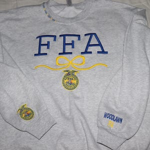 May include: A light gray sweatshirt with blue embroidered "FFA" lettering and a yellow bow design. Below the bow is a circular emblem. The sleeve has a matching emblem and the word "WOODLAWN" in blue.