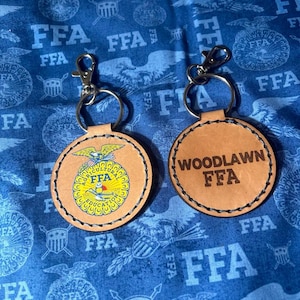 May include: Two leather keychains with silver-colored clasps. One keychain features a yellow and blue FFA emblem with the words "AGRICULTURAL EDUCATION FFA". The other keychain has the words "WOODLAWN FFA" in a brown font.