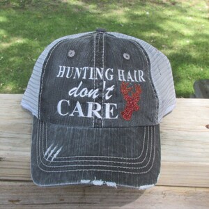 May include: A grey trucker cap with a white mesh back. The front of the cap has the embroidered text "HUNTING HAIR don't CARE" in white, with a red glitter deer head graphic.