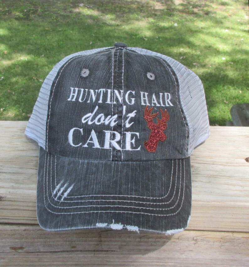 May include: A grey trucker cap with a white mesh back. The front of the cap has the embroidered text "HUNTING HAIR don't CARE" in white, with a red glitter deer head graphic.