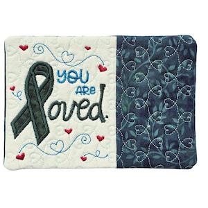 Puede incluir: Un artículo de tela acolchada con las palabras "You are loved" bordadas en azul y negro. Una cinta color turquesa y corazones rojos decoran el lado izquierdo, mientras que el lado derecho presenta un estampado floral azul.