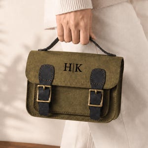May include: An olive green satchel bag featuring a top handle and a front flap secured by two dark charcoal straps with brass-toned metal buckles. The center of the flap is embossed with the black capital letters H|K in a serif font style.