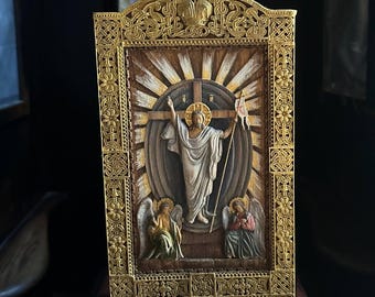 May include: A detailed, wooden religious icon featuring a central figure with outstretched arms, standing before a cross. The icon is framed in gold, with two angels at the bottom. The background has a sunburst design.