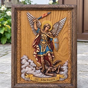 May include: A rectangular wooden wall plaque featuring a relief carving of St. Michael the Archangel with wings, a sword, and a shield, standing over a figure. The piece is framed in dark wood and includes a gold banner with the text St. Michael.