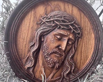May include: A wooden relief carving of a man with long hair, a beard, and a crown of thorns. The carving is set within a circular frame. The wood has a natural grain and a warm, brown color. The background is blurred.
