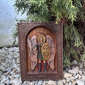 May include: A wooden relief carving of an angel, framed in a dark brown border. The angel is depicted with wings, holding a sword and shield. The carving is set against a backdrop of green foliage and grey rocks.