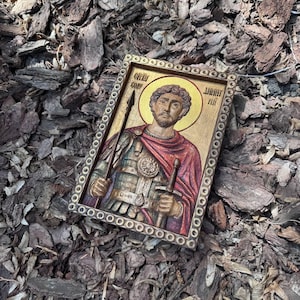May include: A framed religious icon featuring a saintly figure with a golden halo, holding a spear and a sword. The icon has a detailed, carved design with a gold and red color scheme, set against a wooden background. The frame is a dark brown color.