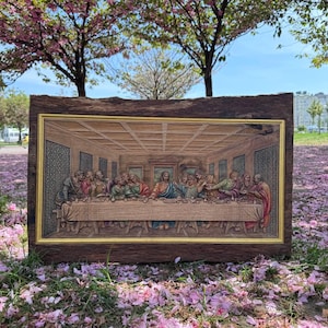 May include: A framed wooden relief carving depicting the Last Supper. The artwork features detailed figures around a long table, with a gold-colored inner frame and a dark brown outer frame. The scene is set indoors, with a focus on the central figures.