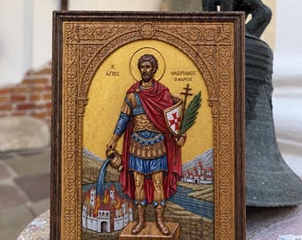 May include: A rectangular wooden icon featuring a relief carving of Saint Florian in armor, holding a vessel pouring water onto a burning building. The figure stands against a gold background within an ornate frame with Greek text inscriptions.