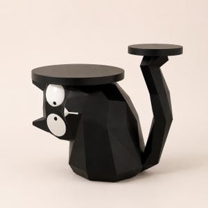 May include: A black cat-shaped side table with a faceted geometric body, two large white circular eyes, and a small white nose. The table features a flat circular top surface and a tall, angular tail that supports a smaller circular platform.